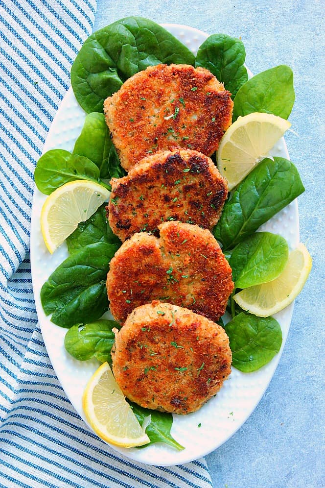 Overhead shot of four tuna cakes on spinach leaves, with lemon slices on the sides, in white oblong serving dish.