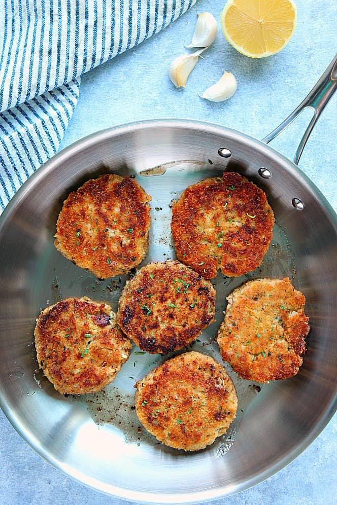 Overhead shot of 6 tuna cakes in a stainless steel pan with striped kitchen towel in background.