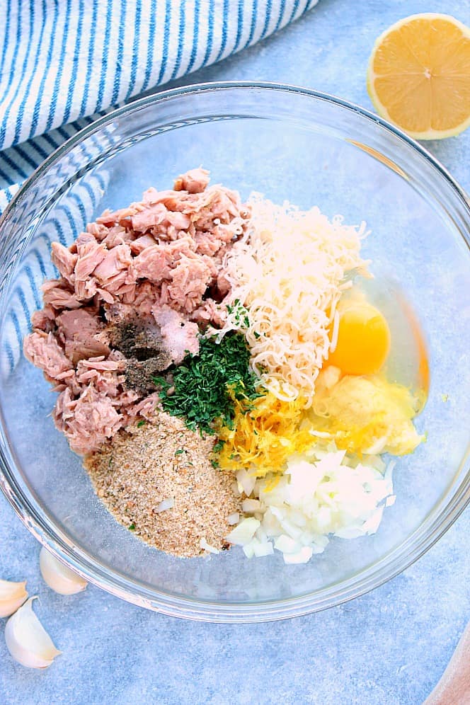 Overhead shot of tuna cakes ingredients: canned tuna, breadcrumbs, egg, cheese, lemon zest, onion and herbs in glass mixing bowl. 