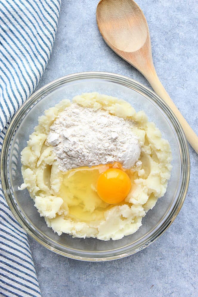 Overhead shot of mashed potatoes, egg and flour in glass bowl.