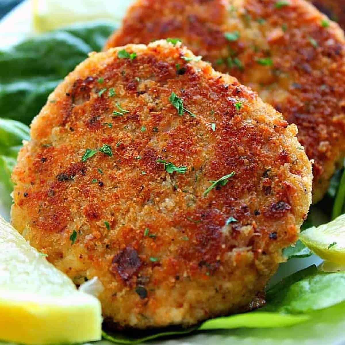 Square image of two tuna cakes on lettuce leaves on white plate.