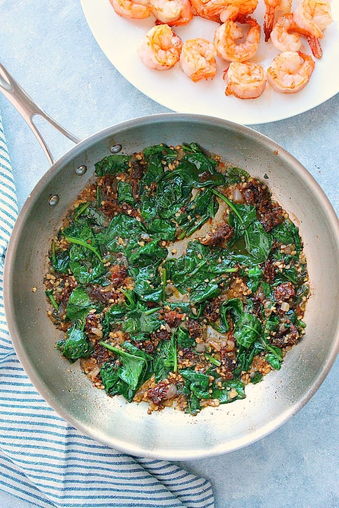 Overhead shot of sauteed spinach and sun-dried tomatoes in stainless steel pan, plate with cooked shrimp next to it.