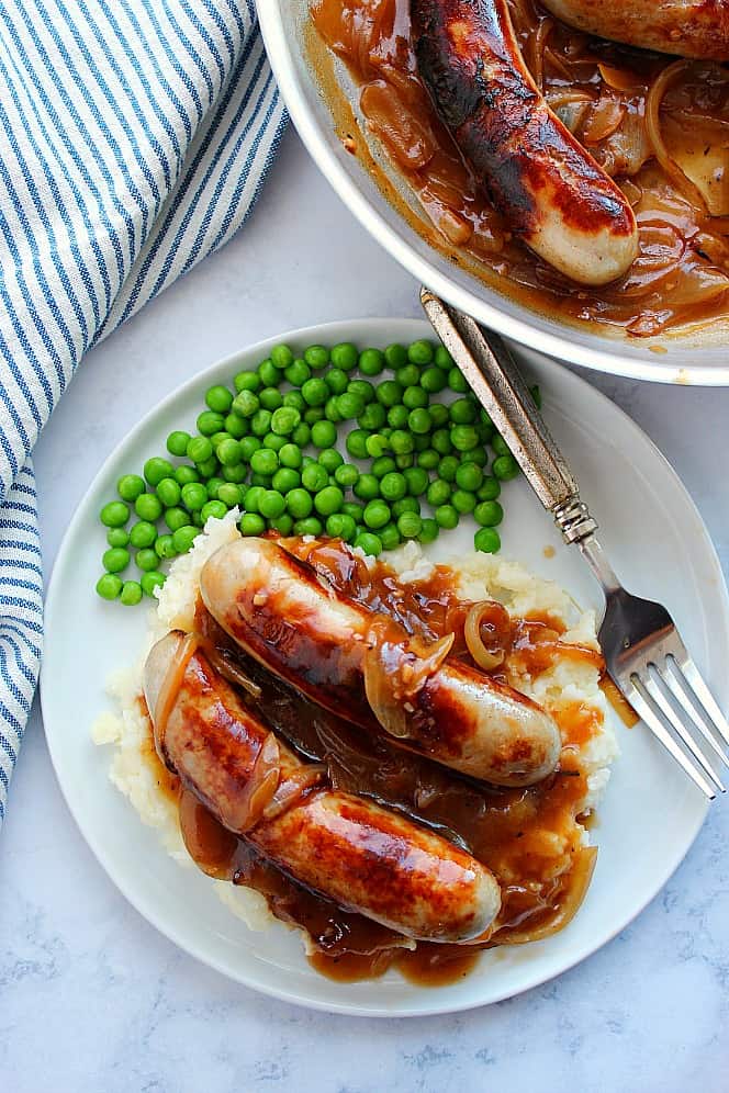 Overhead shot of bangers and mash with peas on white plate and another sausage in the skillet.