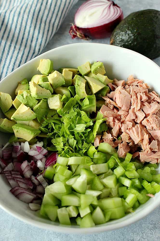 Side shot of tuna, avocado, celery, red onion and cucumber in white salad bowl.
