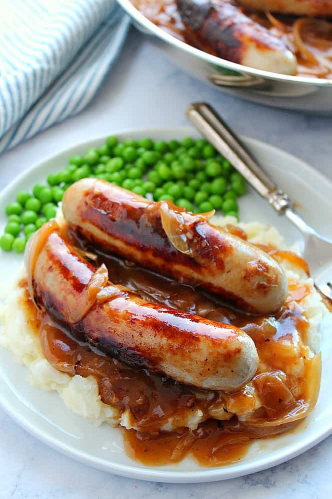 Side shot of bangers and mashed potatoes with onion gravy on white plate, green peas on the side.