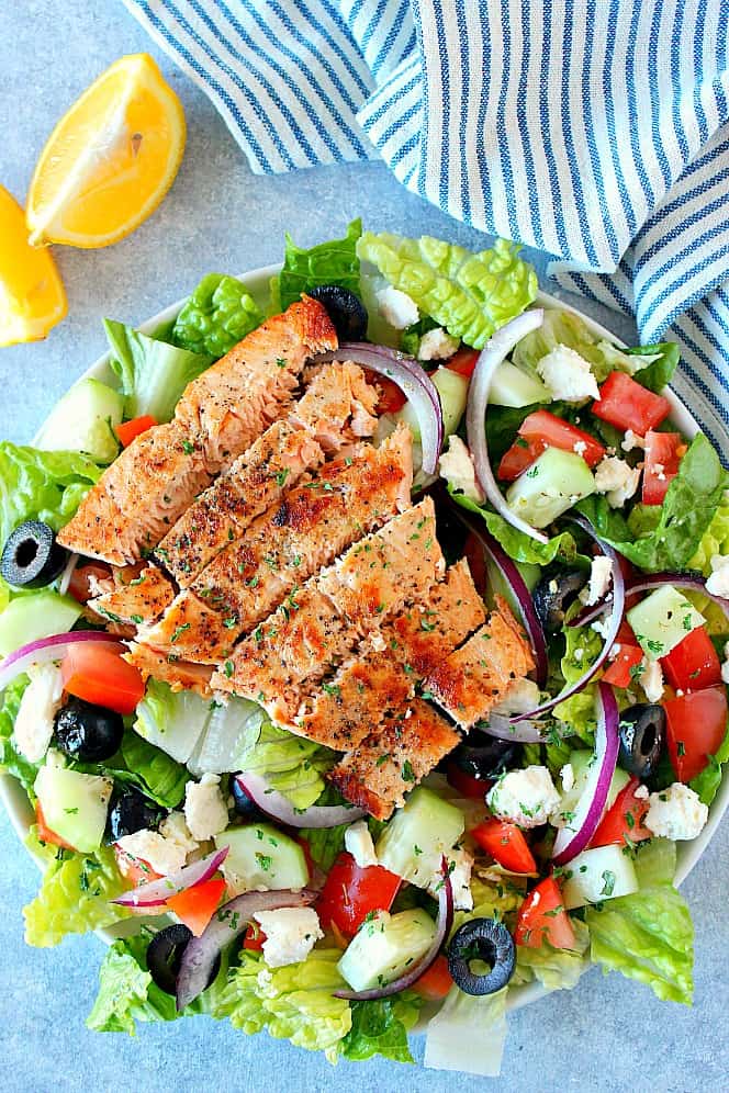 This Greek Salmon Salad is packed with fresh veggies and served with pan-seared salmon. Crisp Romaine lettuce, Roma tomatoes, olives, cucumber, red onion and quick homemade Greek dressing are perfect with fish! Overhead shot of Greek salad with sliced salmon on top, on plate.