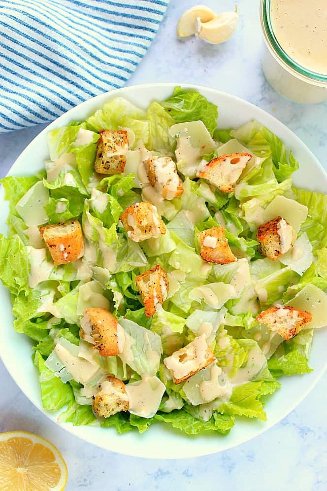 This Caesar Salad is quick, simple and bursting with flavor! You only need Romain lettuce, Parmesan cheese, croutons and dressing to make it! Overhead shot of Caesar salad with croutons on white salad bowl.