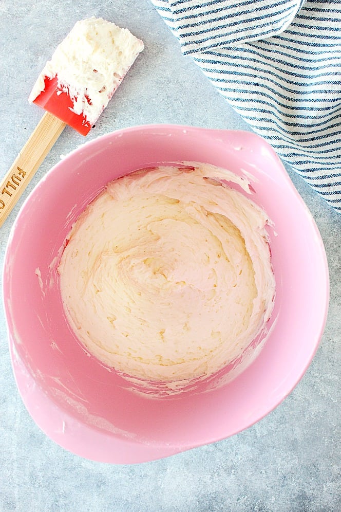 Overhead shot of frosting in pink mixing bowl, more frosting on spatula next to bowl.