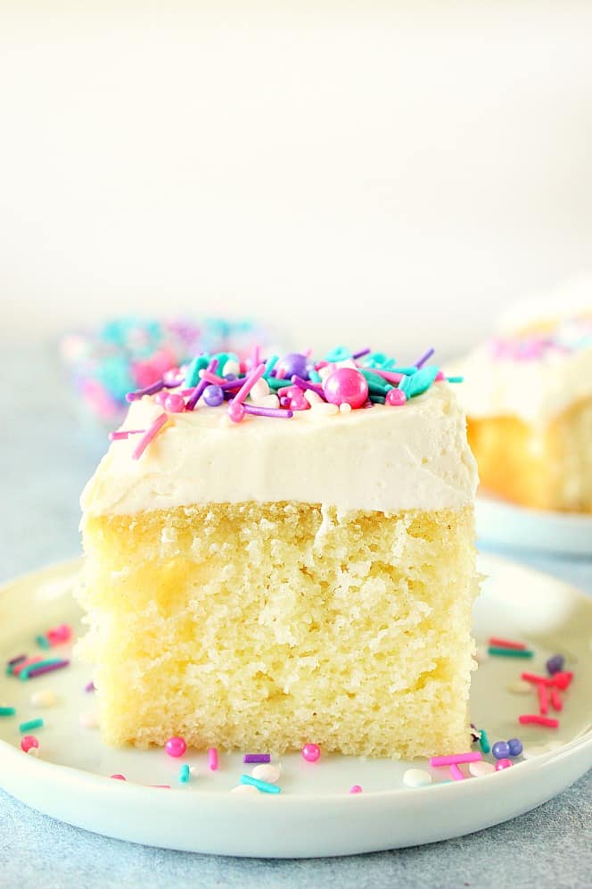 Side shot of a square piece of vanilla cake with white frosting and sprinkles on white plate.