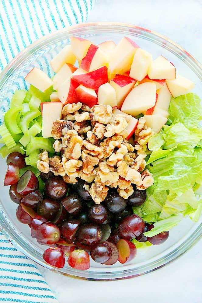 Overhead shot of chopped apples, walnuts, celery, grapes and lettuce in glass mixing bowl.