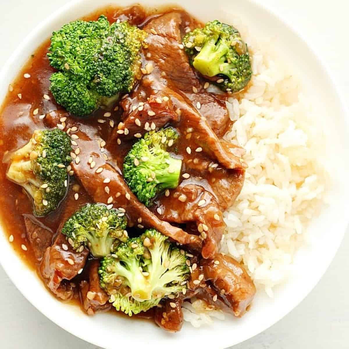 Square image of rice, beef and broccoli in sauce in a white bowl on white wood board.