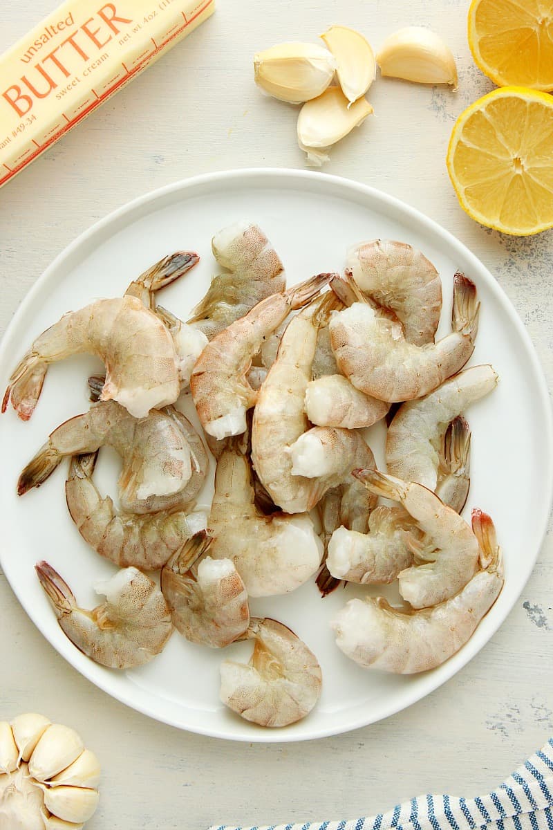 Butter, garlic cloves, lemon and raw shrimp on a plate, set on a gray board.