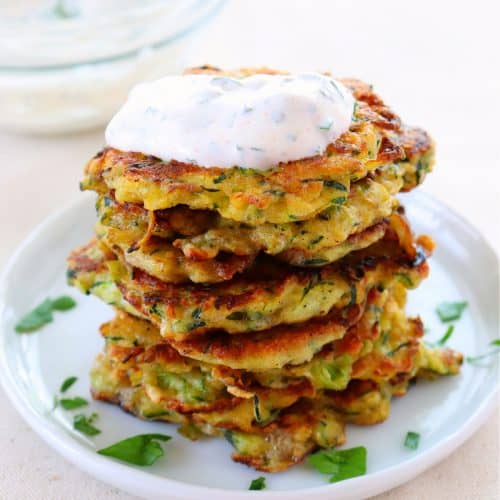 Stack of zucchini fritters with topping on a plate.