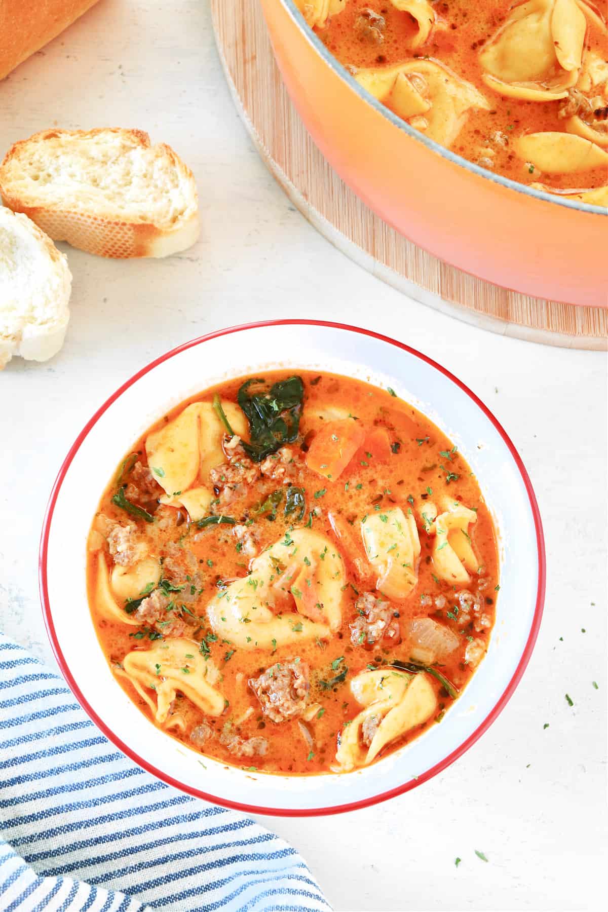 Tortellini soup in a bowl with red rim and pieces of baguette next to it.