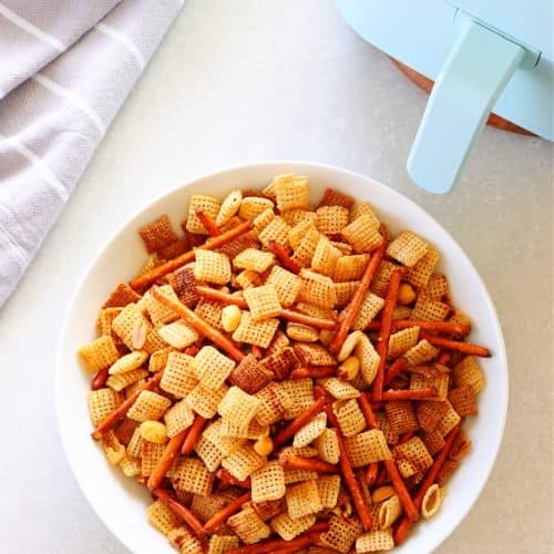 Party mix in white bowl next to air fryer.