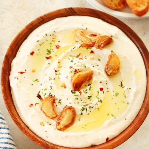 Feta dip with roasted garlic cloves in a wooden bowl on a cream board.