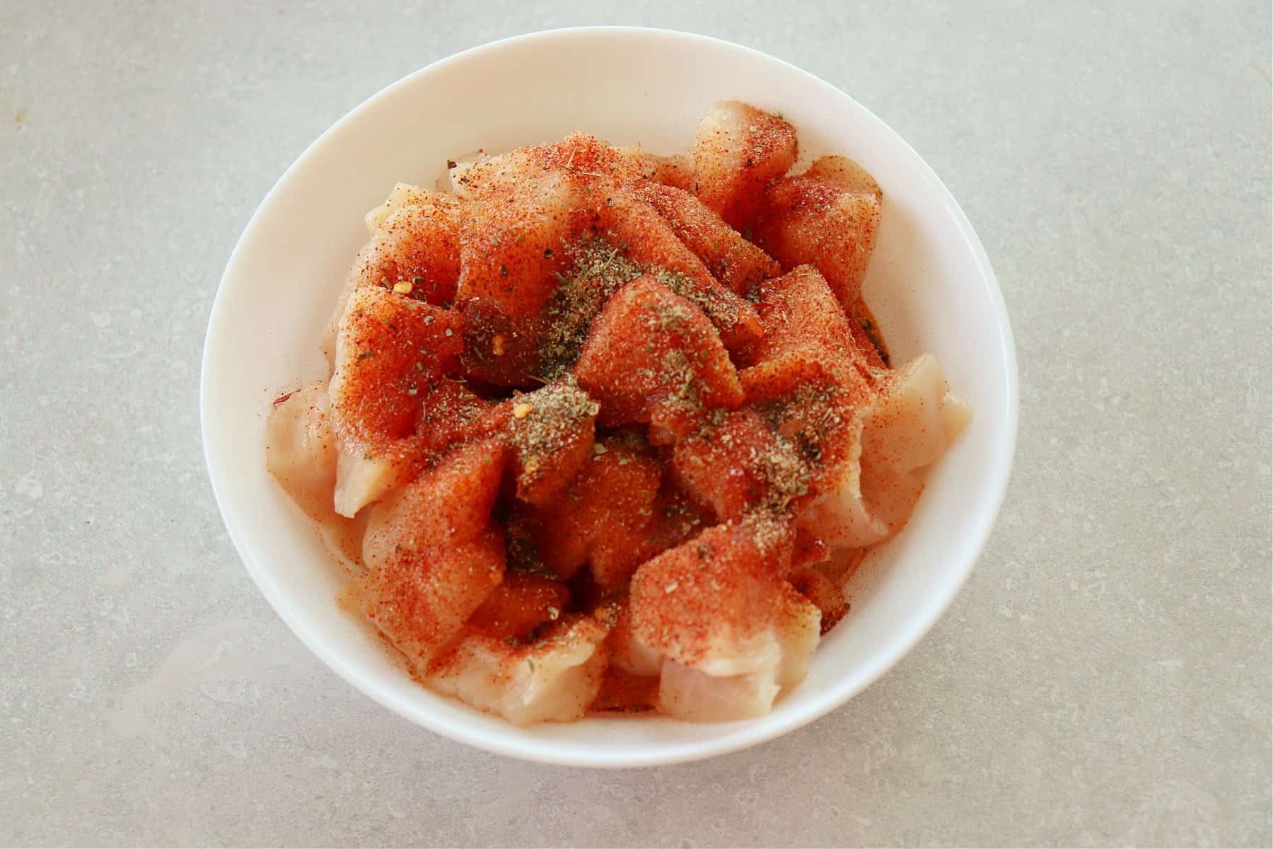 Raw chicken pieces with seasoning in a white bowl.