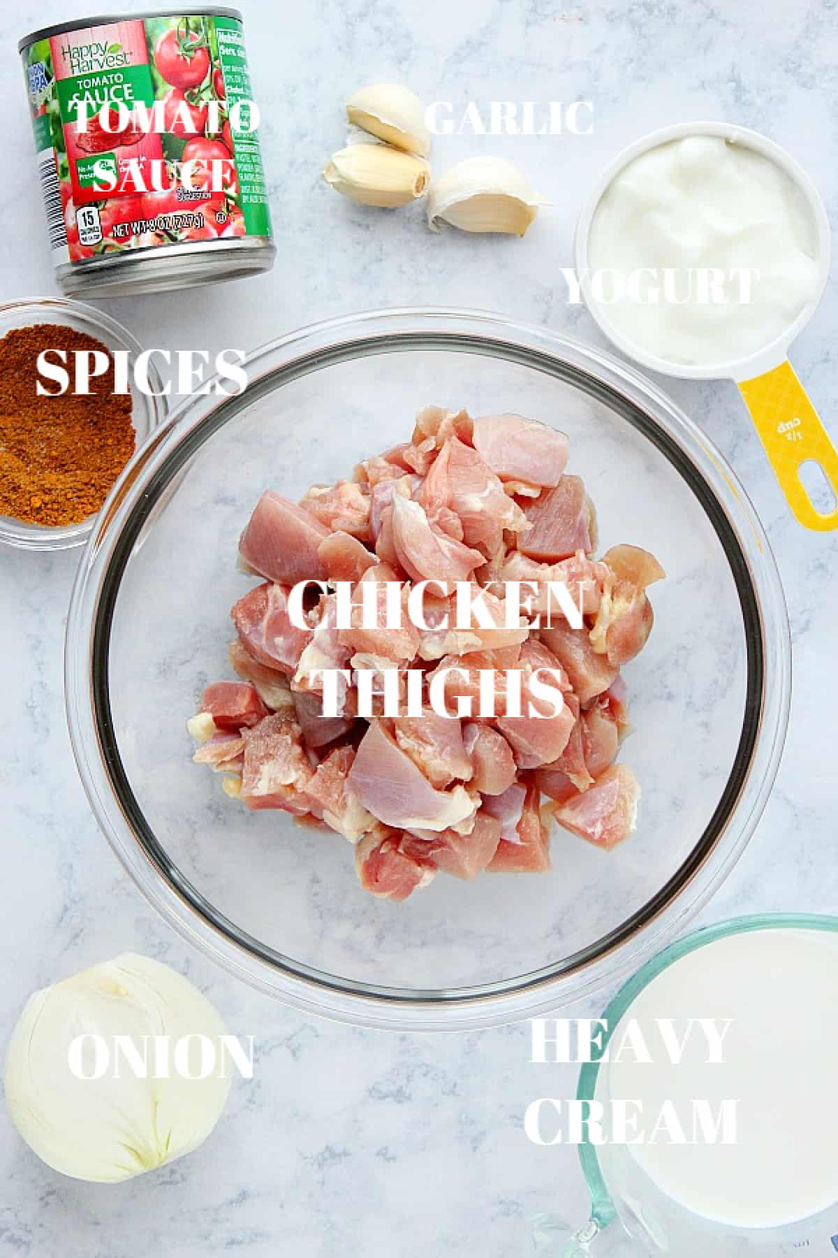 Chicken thighs pieces in glass bowl, onion, heavy cream, garlic, tomato sauce in a can, spices in a bowl and yogurt on a gray board.