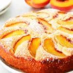 Side shot of cake with peach slices and powdered sugar on a white plate with fresh peaches behind it.