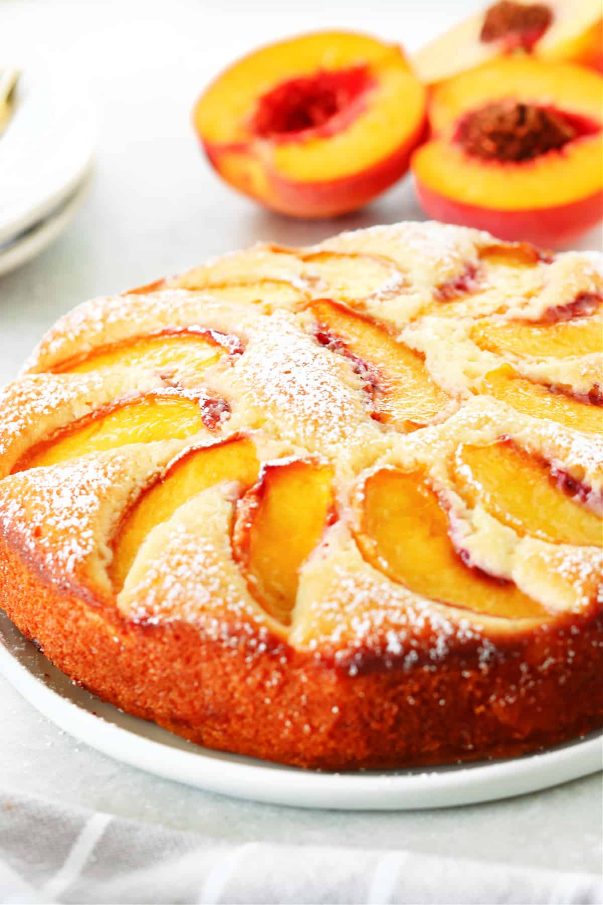 Side shot of cake with peach slices and powdered sugar on a white plate with fresh peaches behind it.