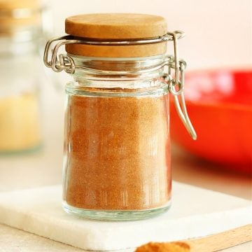 Pumpkin pie spice mix in a small spice jar with hinge top, set on a marble coaster, with a wooden spoon infront of it.