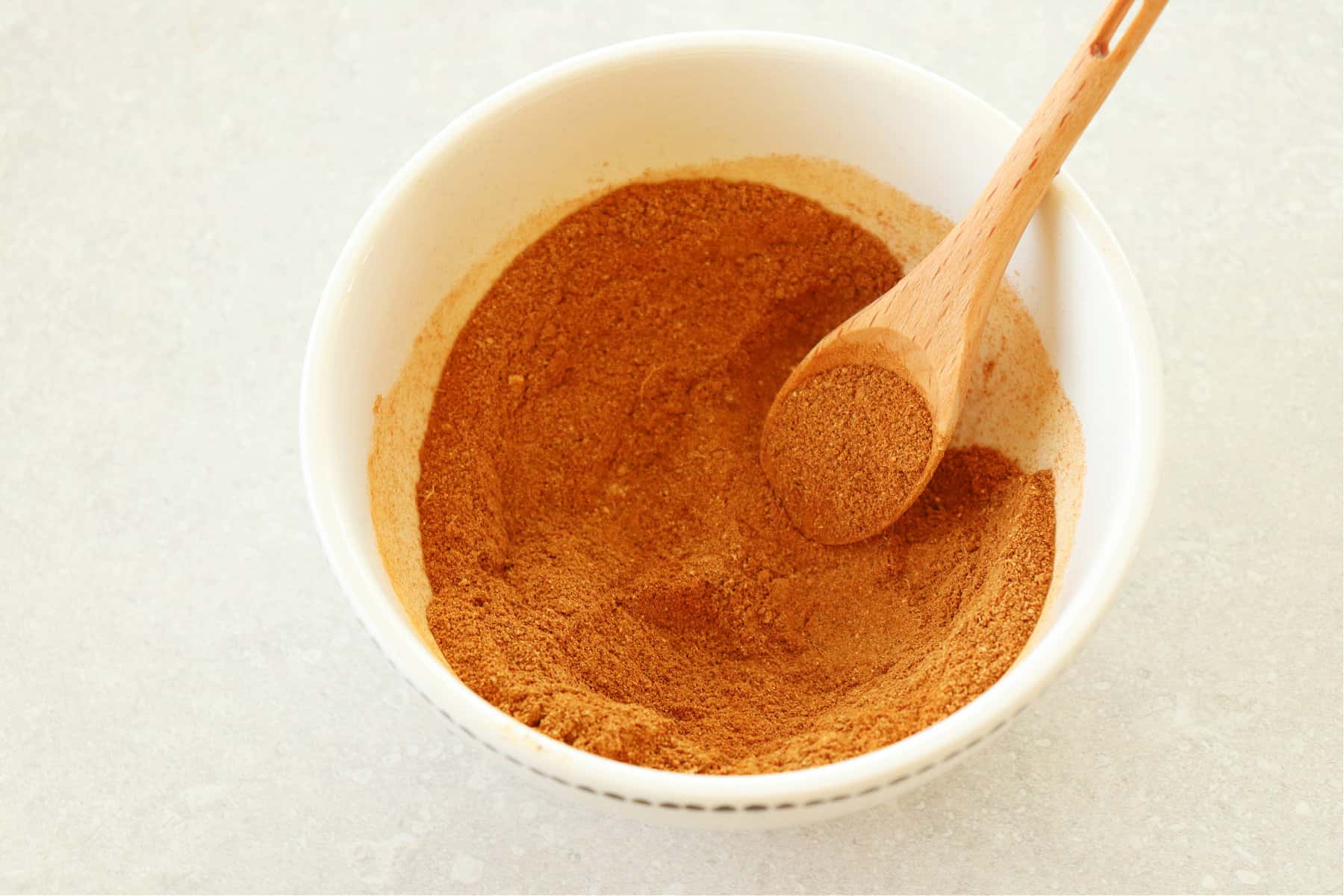Pumpkin pie spice mix in a small white bowl with a wooden measuring spoon, on a gray board.