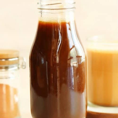Square photo of pumpkin syrup in a glass bottle with a mug with coffee in the background.