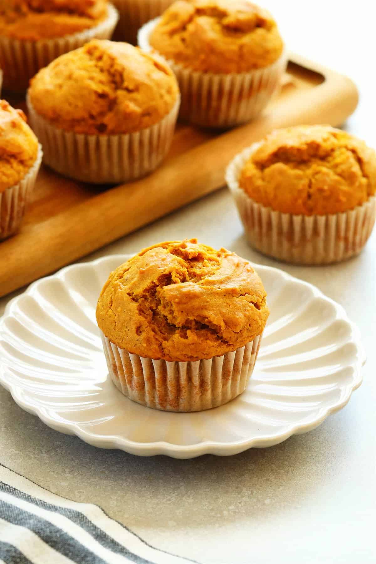 Pumpkin muffin on a dessert plate, one muffin next to the plate and many muffins on a wooden cutting board in the background.