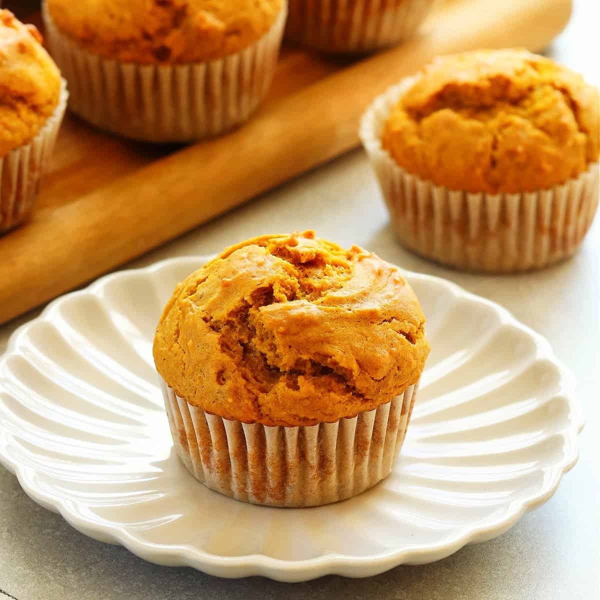 Square photo of a pumpkin muffin on a petal plate with more muffins in the background.