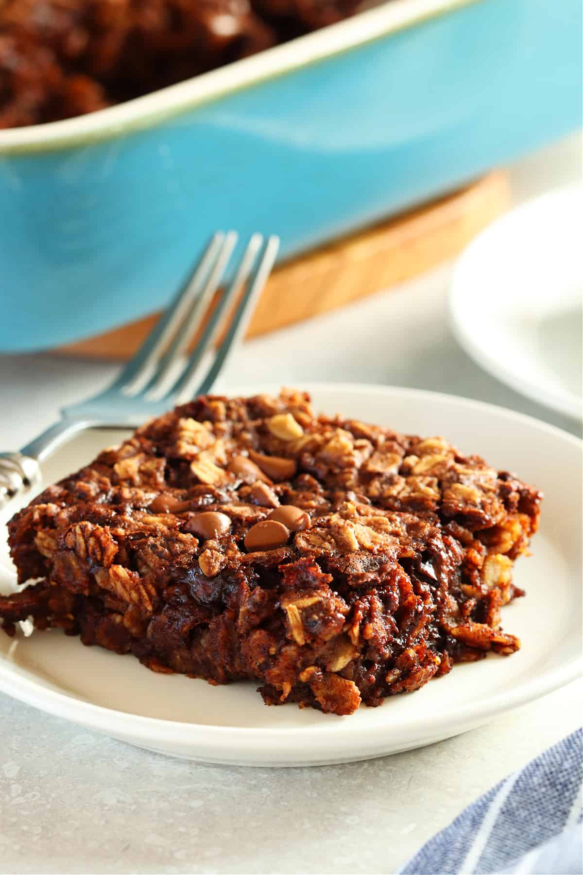 One serving of baked chocolate oatmeal on a small white plate with the baking dish in the background.