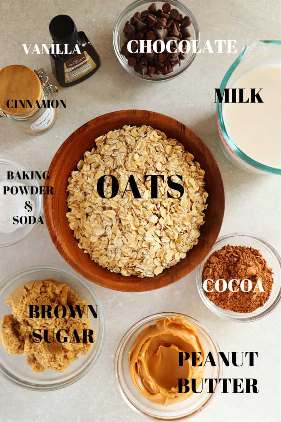 Oats, brown sugar, peanut butter, cocoa powder, milk, cinnamon, vanilla, baking powder and soda in glass containers on a gray board.