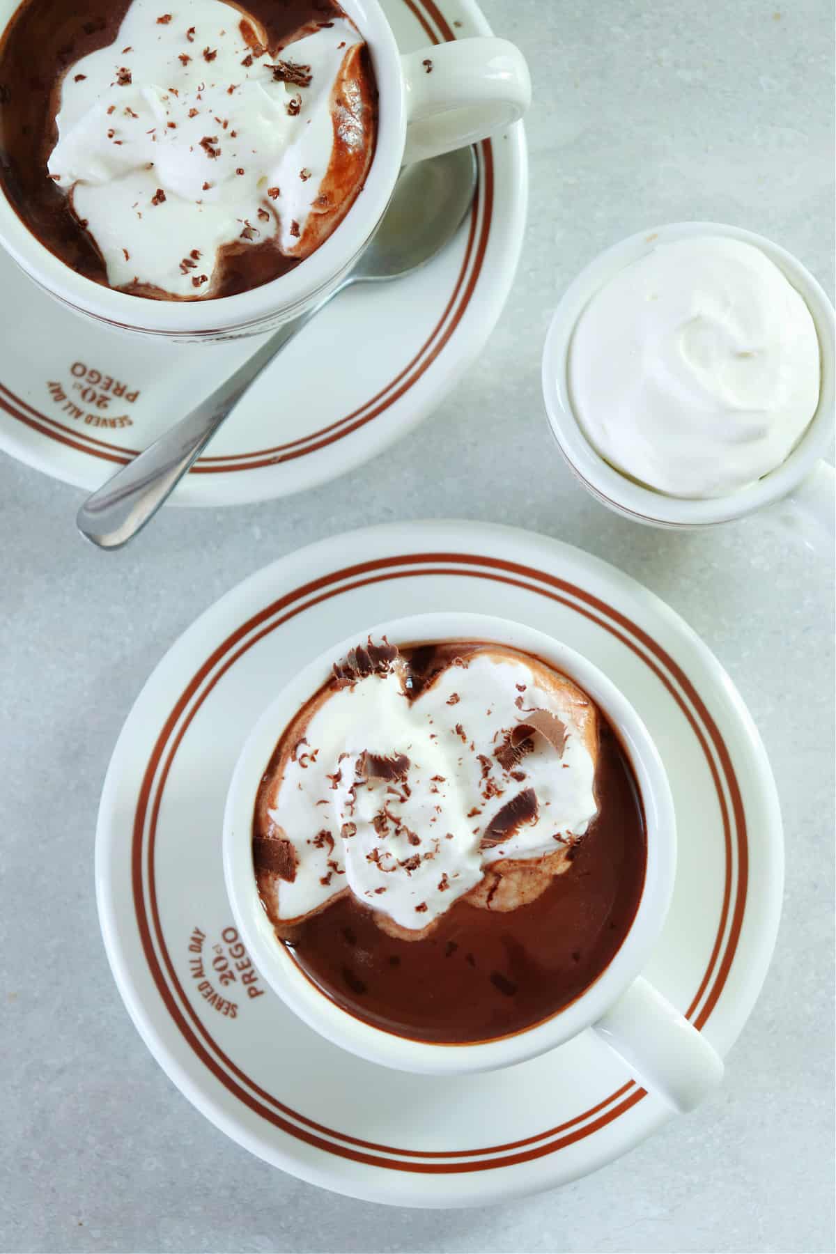 Overhead photo of hot chocolate in two mugs on saucers set on a gray board.