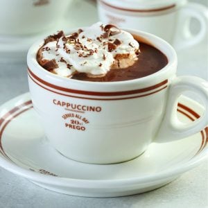 Square image of hot chocolate in a white mug on a saucer, with whipped cream and shaved chocolate on top.