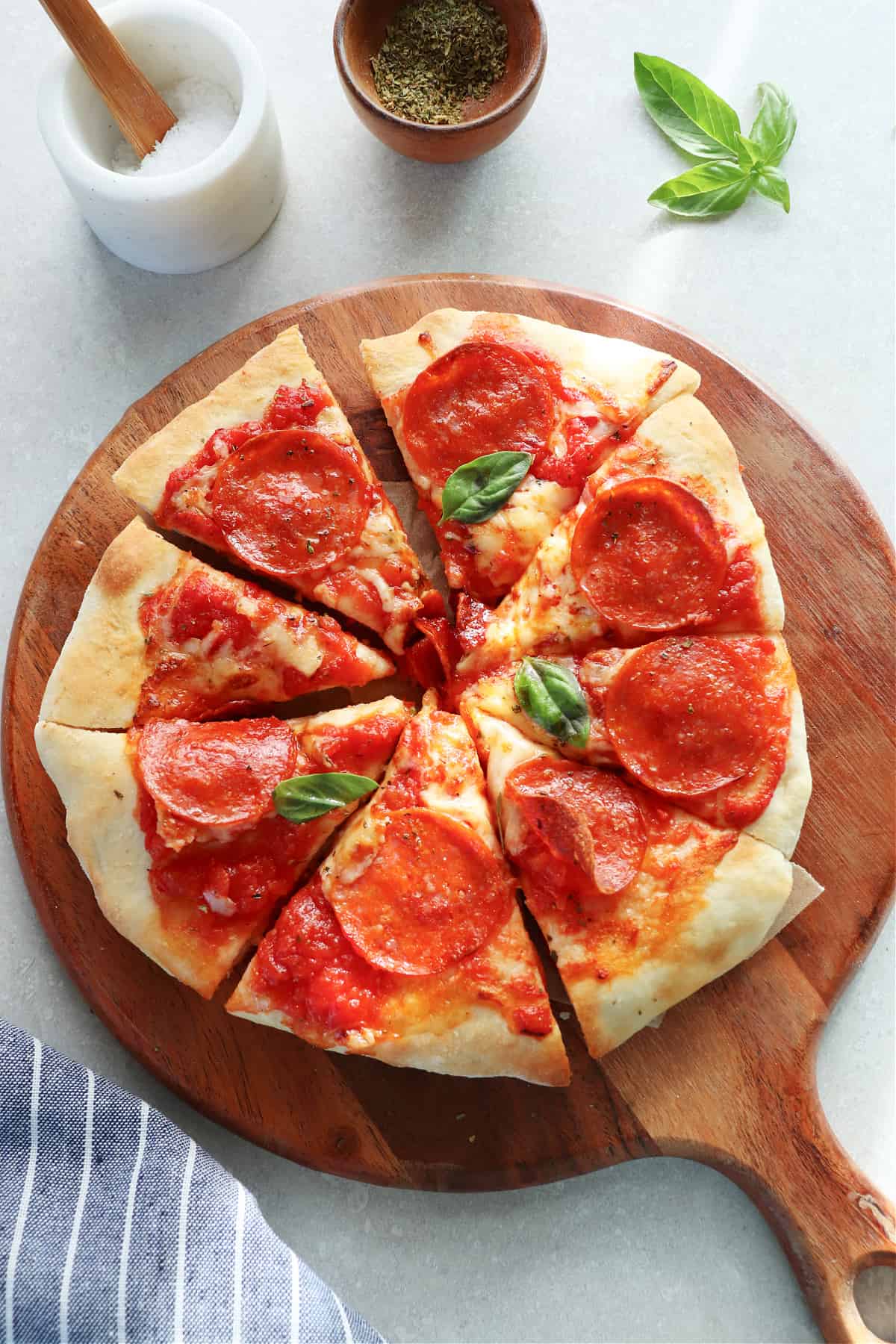 Sliced pizza on wooden board, basil leaves, salt and seasoning in small cups next to it.