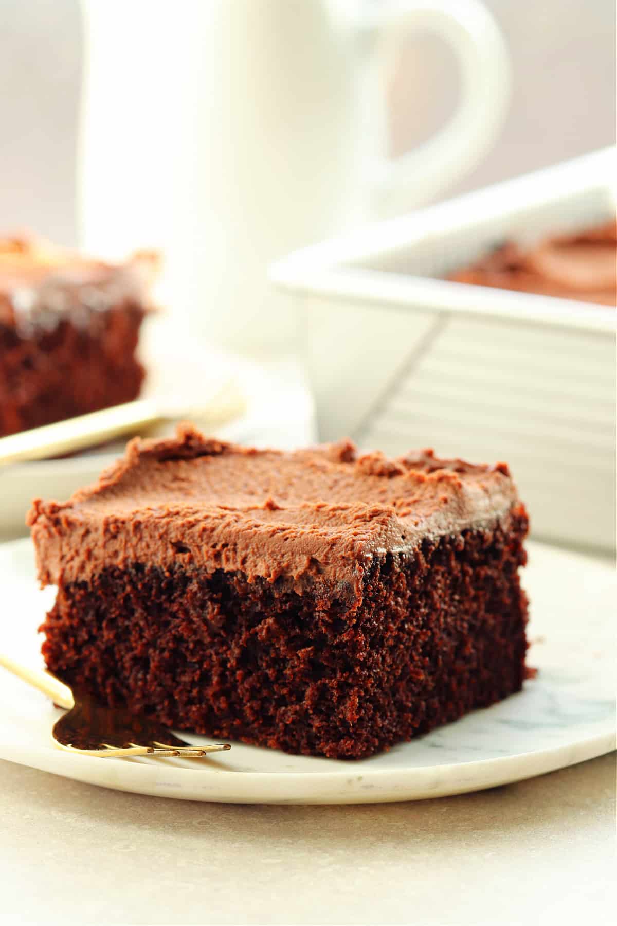 One piece of chocolate cake with frosting on a white plate with a pan with the cake in the background.