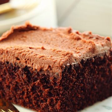 A piece of chocolate cake with chocolate frosting on a marble plate with gold fork.