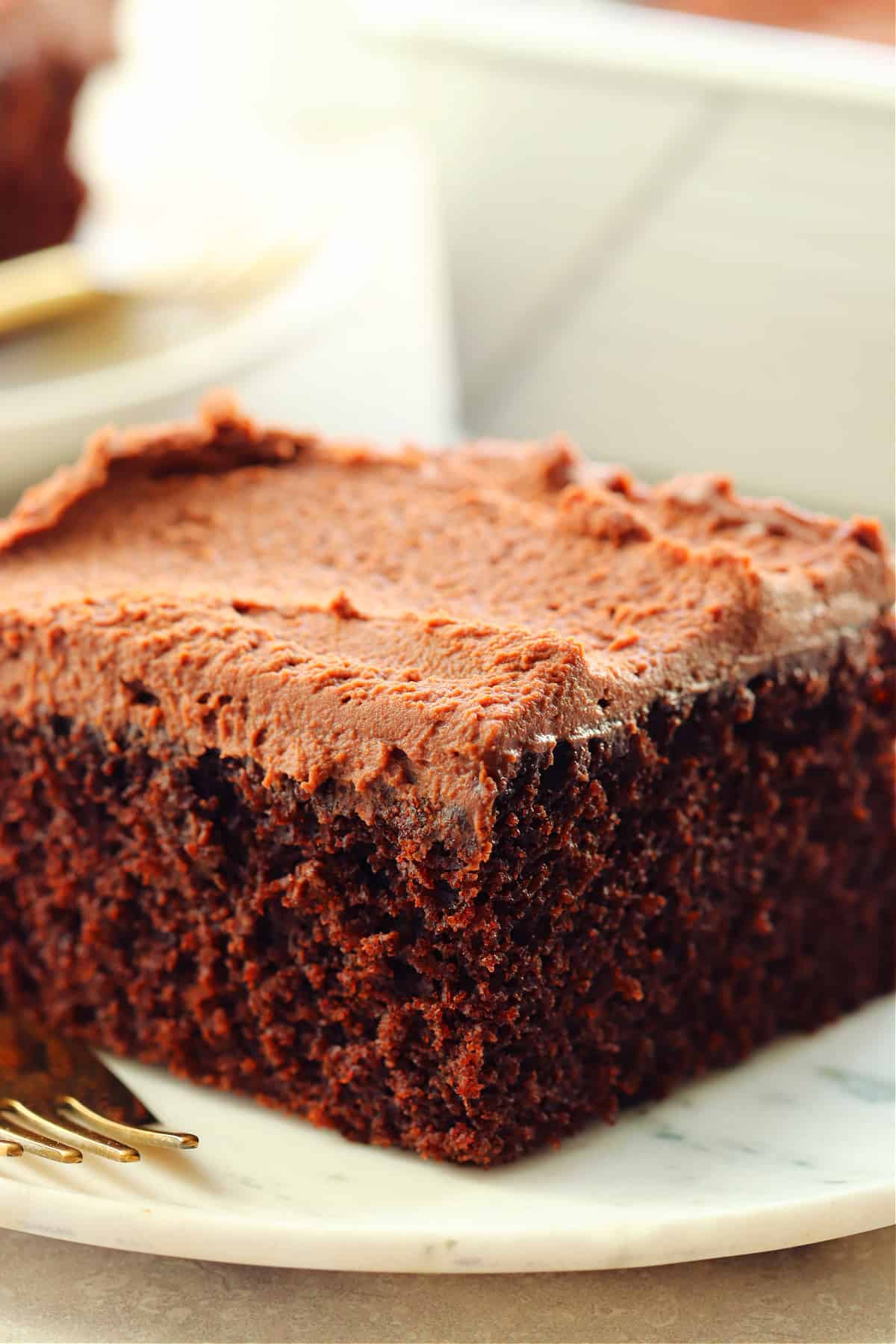 A piece of chocolate cake with chocolate frosting on a marble plate with gold fork.