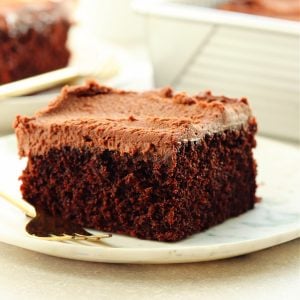 Square image of a serving of eggless chocolate cake with frosting on a white marble plate.