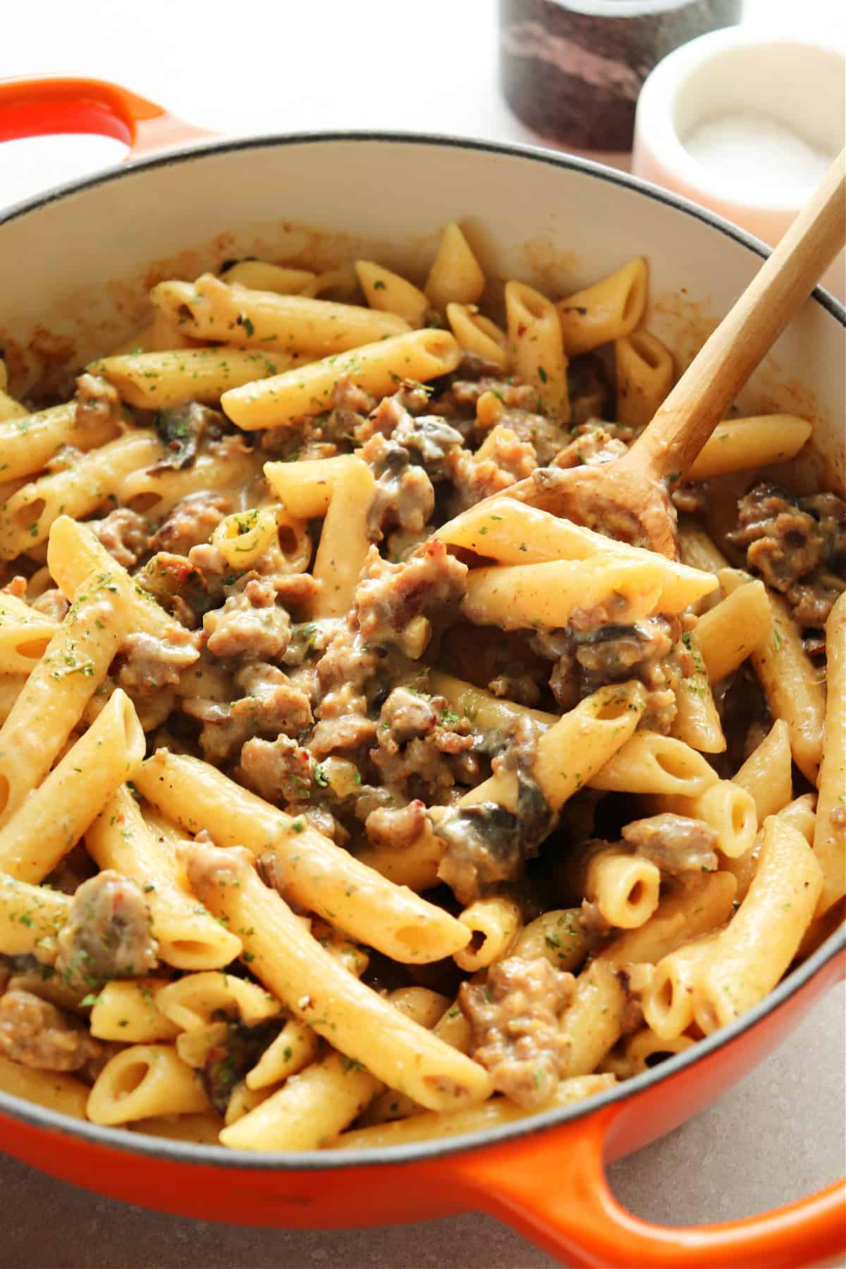 Penne and sausage in an orange Dutch oven on a gray board.