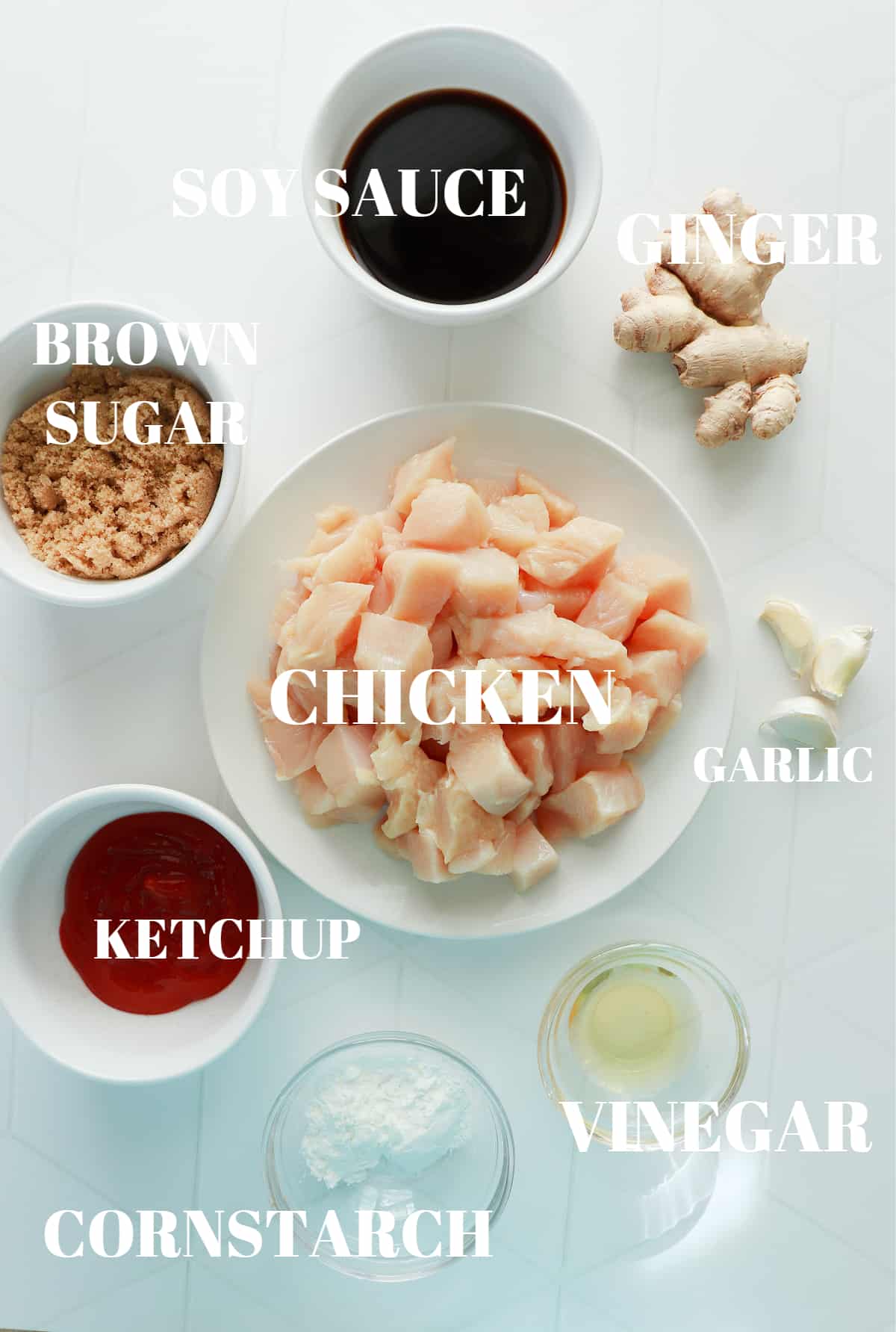 Chicken, soy sauce, ginger, garlic, cornstarch, vinegar, ketchup and brown sugar in bowls on a white tile board.