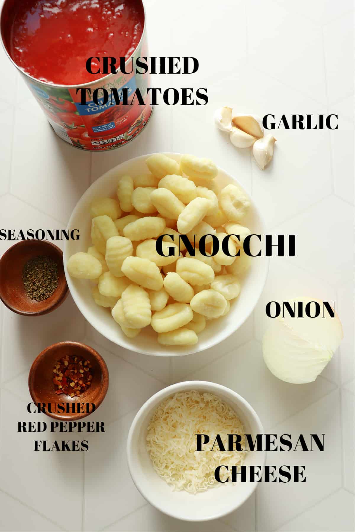 Gnocchi, crushed tomatoes, Parmesan, onion, garlic cloves and seasoning in bowls on a white tile board.