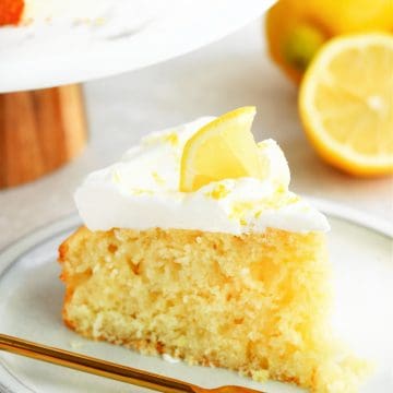One slice of lemon cake with whipped cream and a small slice of lemon on a dessert plate with gold fork.