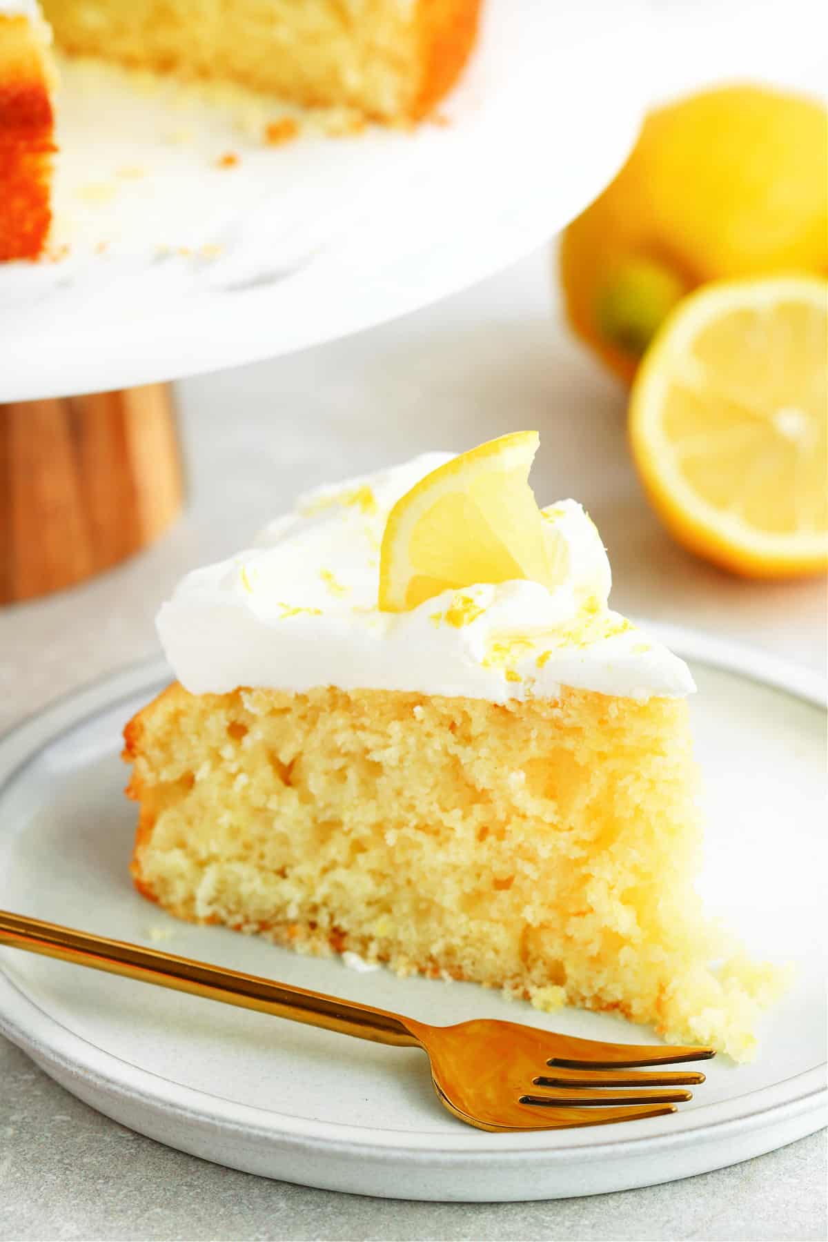 One slice of lemon cake with whipped cream and a small slice of lemon on a dessert plate with gold fork.