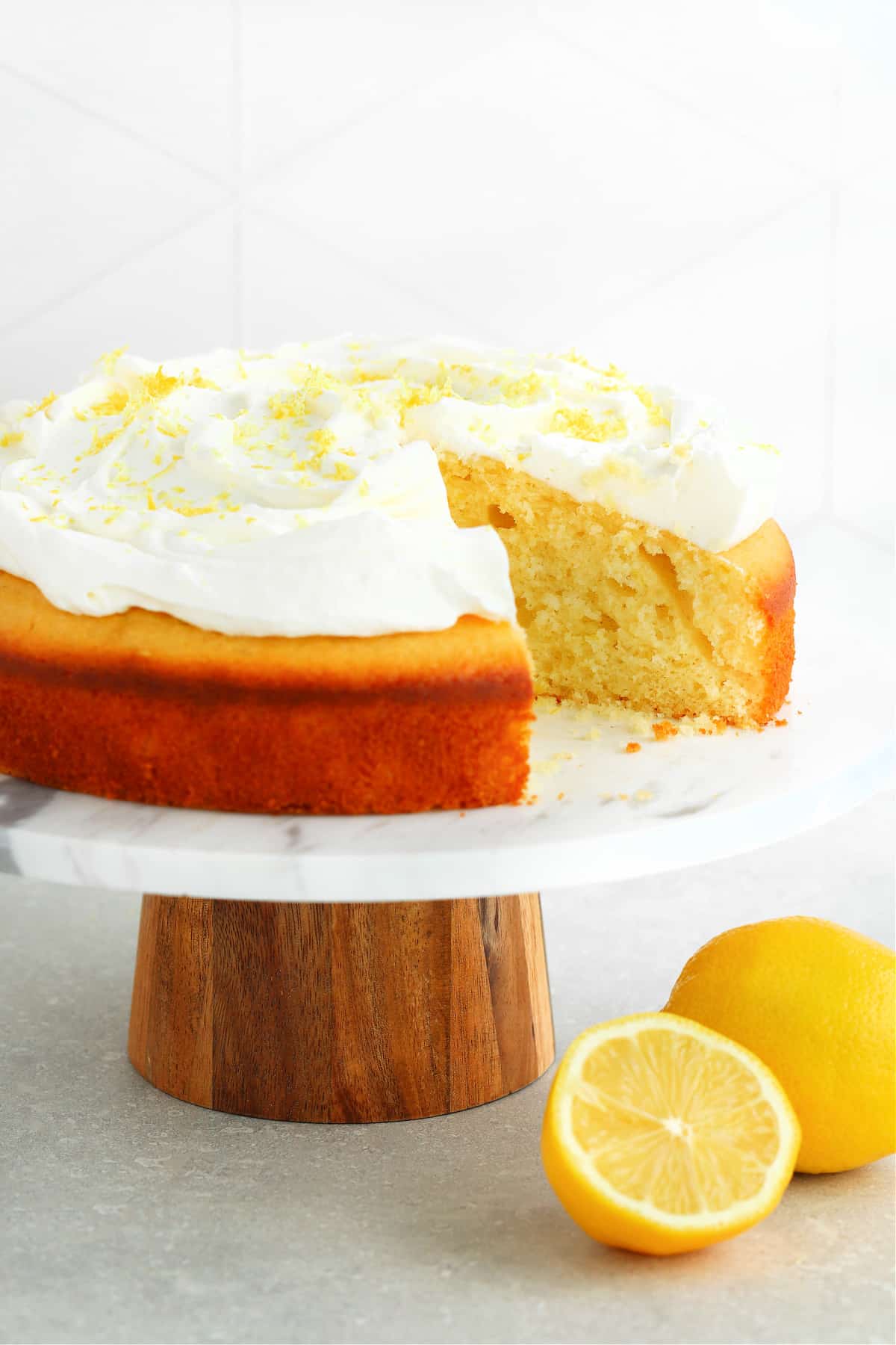 Round lemon cake with whipped cream on a marble cake stand with lemons next to it.