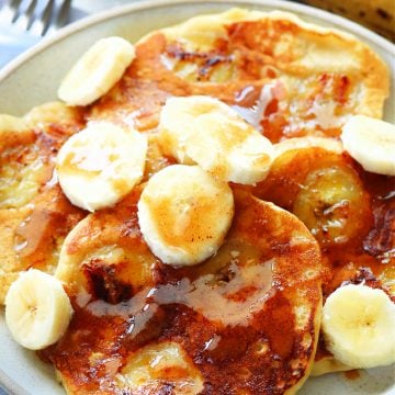 Four banana pancakes, drizzled with brown sugar butter and topped with slices of fresh bananas on a plate with fork.