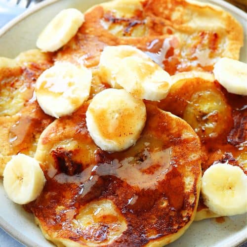 Square image of banana pancakes with slices of fresh bananas and syrup on a plate.
