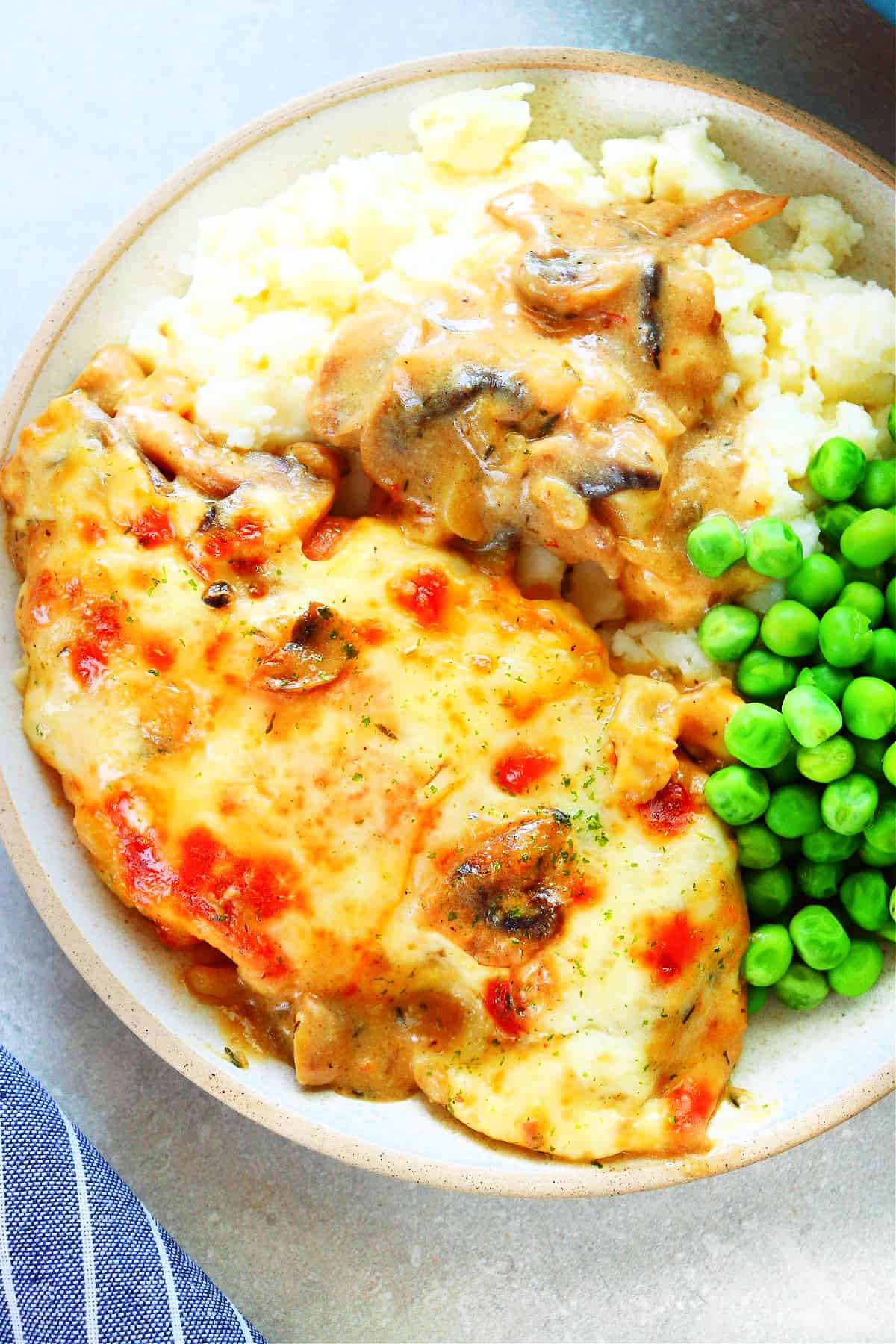 Chicken breast with melted cheese, mashed potatoes with mushroom gravy and steamed untried peas on a dinner plate.