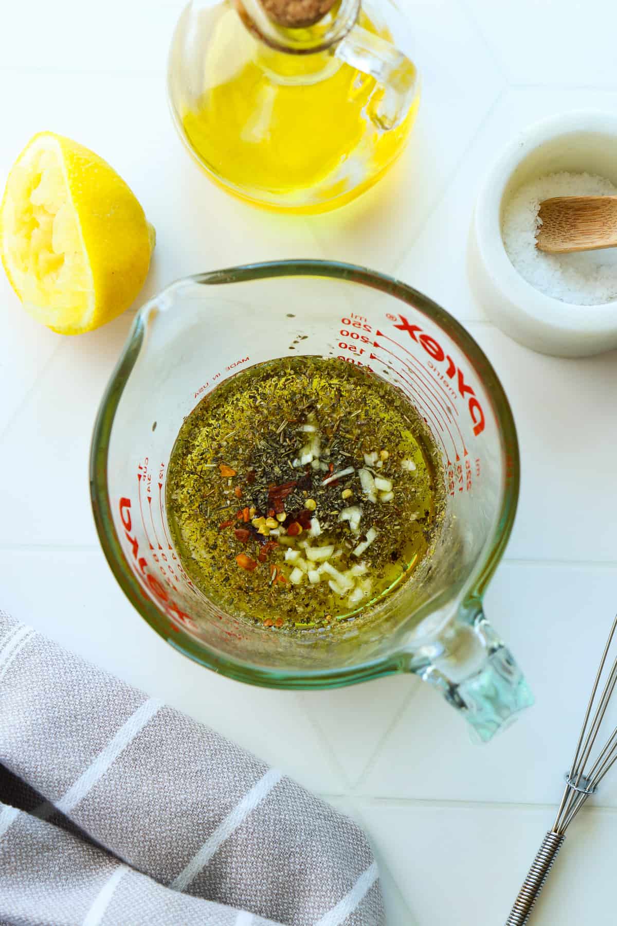 Ingredients for Italian dressing in a glass Pyrex cup with a small whisk next to it and a bottle of olive oil behind it.