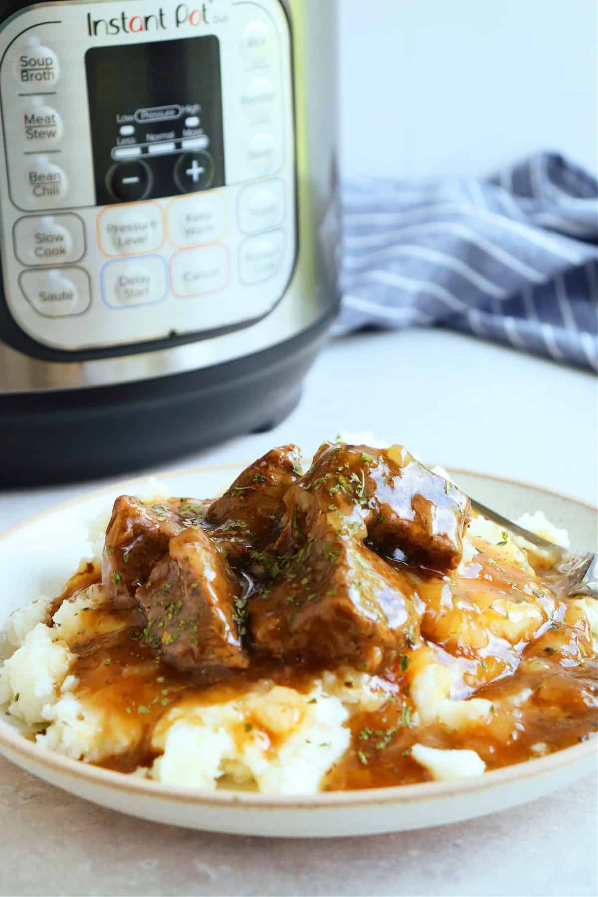 Side photo of beef tips with gravy on mashed potatoes. on a plate, with the Instant Pot in the background.