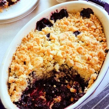 Blueberry crumble in a blue stoneware baking dish with a serving missing, with blue stripped towel next to it, and blueberries in a bowl behind it.